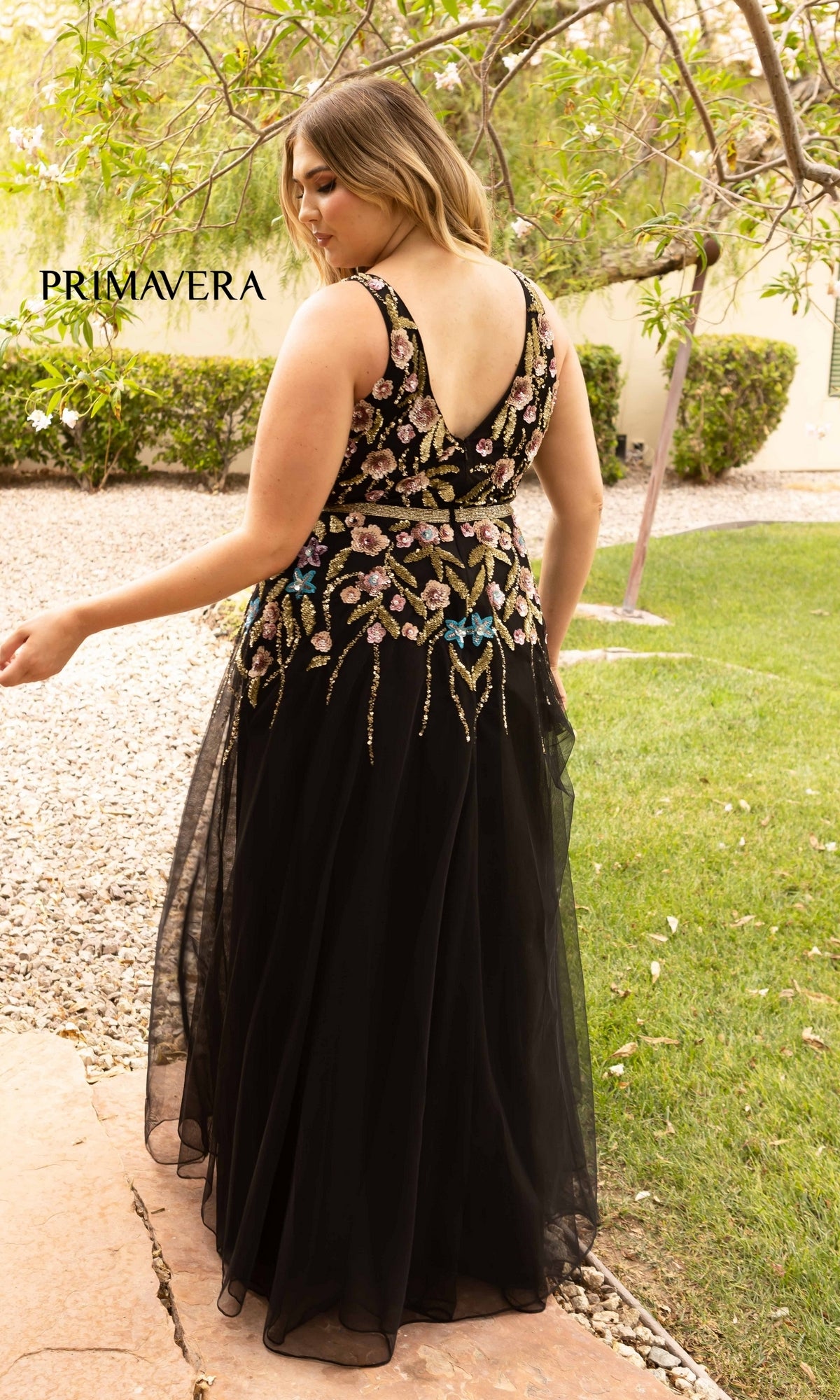 A woman faces away outdoors on a stone path, wearing the Floral-Beaded Long Black Plus-Size Prom Dress with colorful floral embroidery.